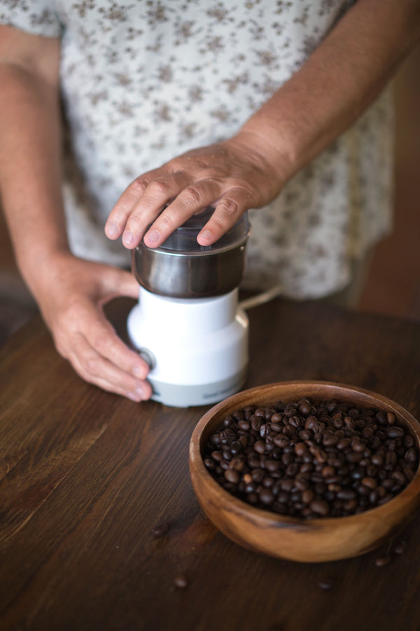Person grinding coffee with a small white electric coffee grinder beside a wooden bowl of beans