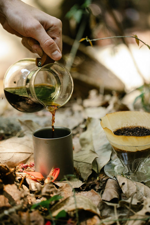 Pouring coffee from a glass carafe into a camping mug beside a glass pour-over dripper outdoors