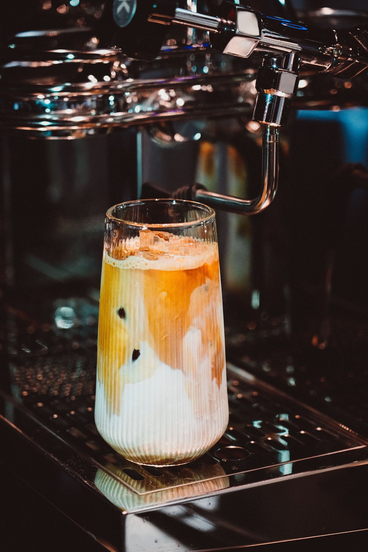 Iced latte in a ribbed glass with espresso and milk swirls on the drip tray of an espresso machine