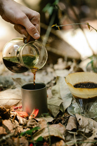 Pouring coffee from a glass carafe into a camping mug beside a glass pour-over dripper outdoors