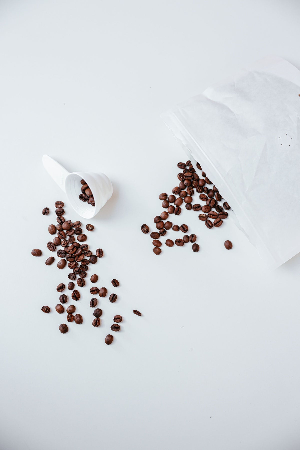 Whole coffee beans spilling from a paper bag beside a white scoop for single dosing coffee beans