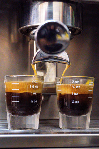 Two espresso shots pouring into glass measuring cups marked in ounces from a split spout